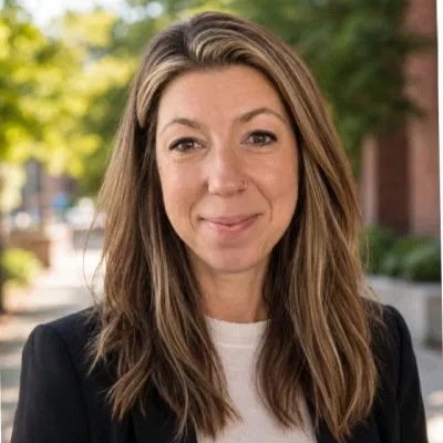 Lizzie Camstra Hughes, smiling, in a dark blazer with trees behind her.